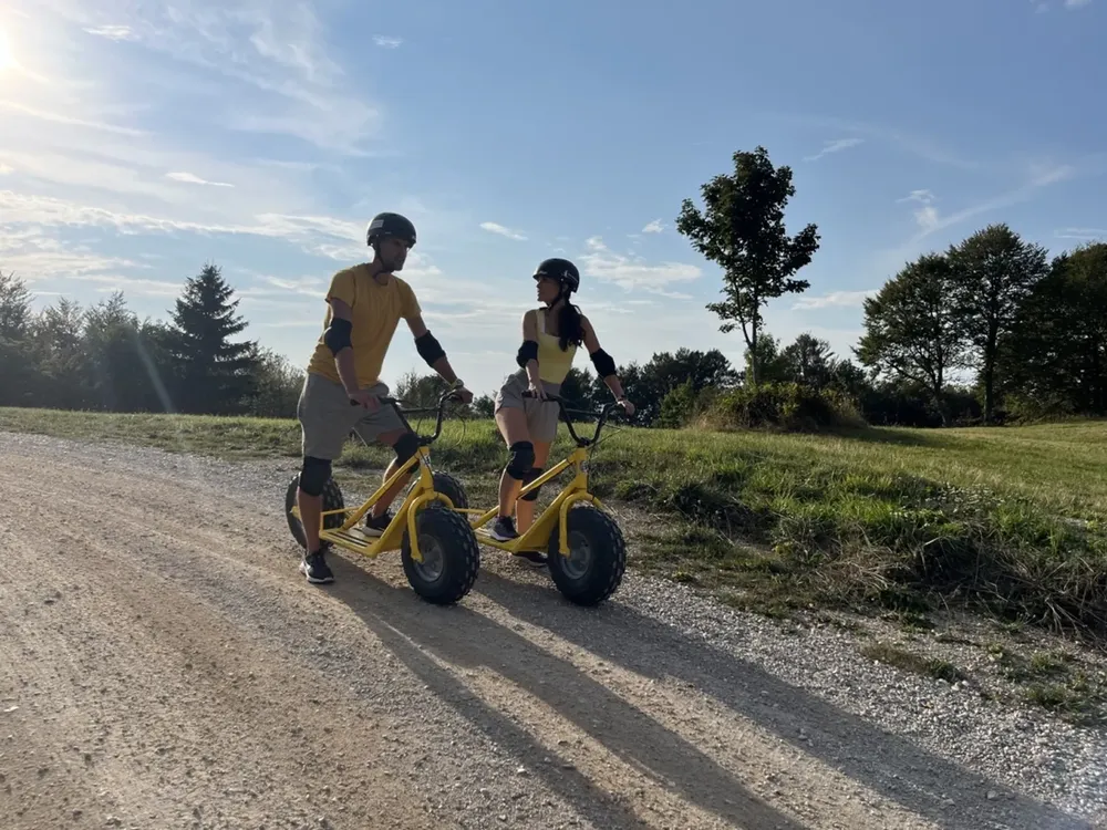 Dve osebi se spuščata z gorskimi skiroji po makadamski poti z vrha Slivnice nad Cerkniškim jezerom, obdani z gozdom in razgledi na notranjsko pokrajino.