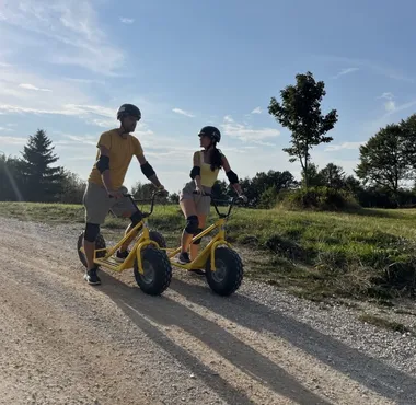 Dve osebi se spuščata z gorskimi skiroji po makadamski poti z vrha Slivnice nad Cerkniškim jezerom, obdani z gozdom in razgledi na notranjsko pokrajino.