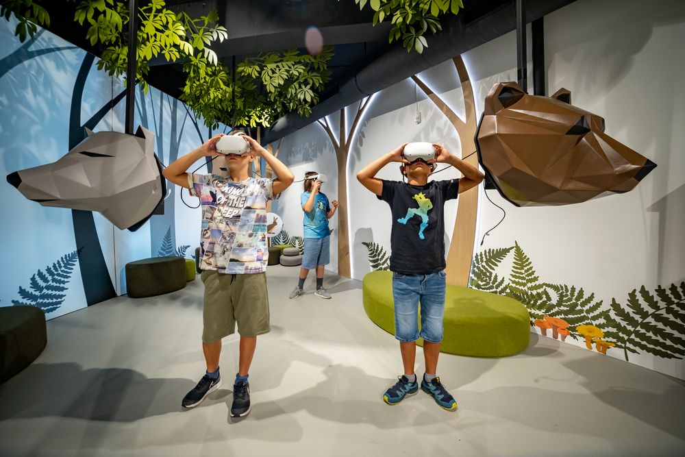 Trije otroci z VR-očali raziskujejo virtualni gozd v interaktivnem razstavnem prostoru. Ob njih visijo stilizirane skulpture volka in medveda, prostor pa krasijo drevesa in gozdni motivi.
